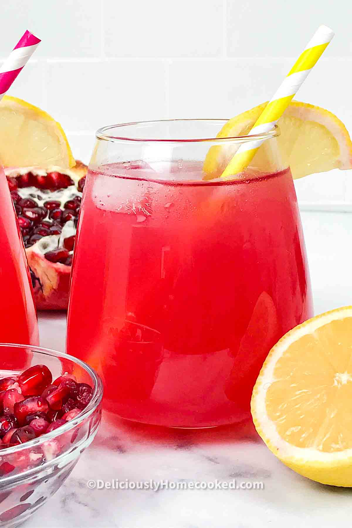 A glass of red juice with a yellow straw in it, lemon slices for garnishing and some open faced pomegranates and a small glass bowl filled with pomegranate seeds 