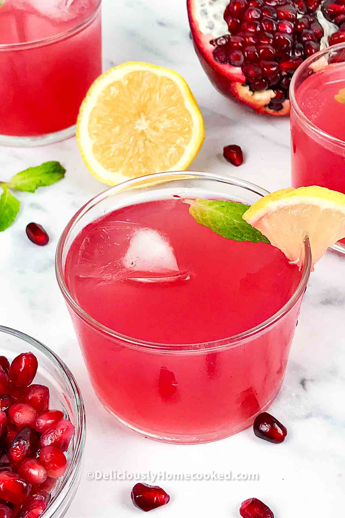 A short glass of pomegranate lemonade with a mint leaf in it, and in the table, lemon slices for garnishing, an open faced pomegranate and a small glass bowl filled with pomegranate arils.