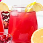 A glass of red juice with a yellow straw in it, lemon slices for garnishing and some open faced pomegranates and a small glass bowl filled with pomegranate seeds
