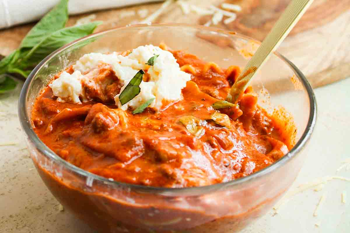 A bowl of creamy tomato basil soup garnished with dollops of ricotta cheese and fresh basil leaves, with a spoon resting inside. The soup is placed on a wooden board with scattered cheese shavings around.